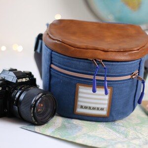 K&ouml;nnte beinhalten: Eine blaue Jeans-Kameratasche mit einer braunen Lederklappe und einem Rei&szlig;verschluss. Die Tasche hat einen wei&szlig;-blau gestreiften Aufn&auml;her mit dem Text "Blah Blah Blah". Im Vordergrund befindet sich eine schwarze Kamera.