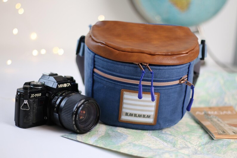 K&ouml;nnte beinhalten: Eine blaue Jeans-Kameratasche mit einer braunen Lederklappe und einem Rei&szlig;verschluss. Die Tasche hat einen wei&szlig;-blau gestreiften Aufn&auml;her mit dem Text "Blah Blah Blah". Im Vordergrund befindet sich eine schwarze Kamera.