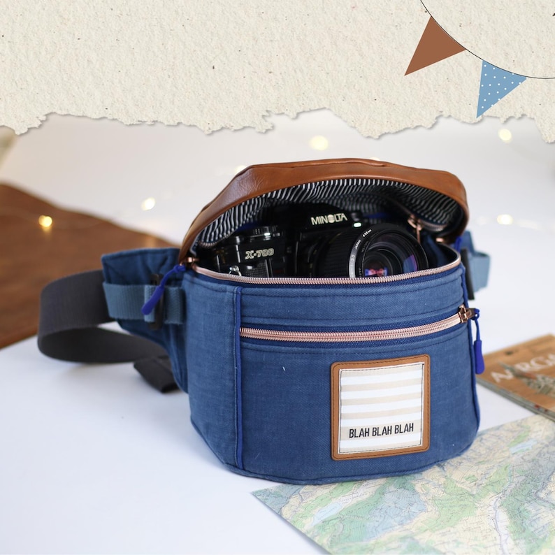 K&ouml;nnte beinhalten: Eine blaue Jeans-Bauchtasche mit einer braunen Lederklappe und einem Rei&szlig;verschluss. Die Bauchtasche hat einen wei&szlig;-beigen Streifen-Patch mit dem Text "Blah Blah Blah". Eine schwarze Kamera befindet sich in der Bauchtasche.