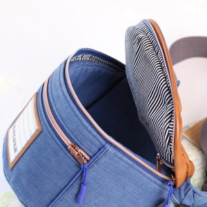 K&ouml;nnte beinhalten: Eine blaue Jeans-Bauchtasche mit einem braunen Lederriemen und einem Rei&szlig;verschluss. Das Innere der Tasche ist mit schwarz-wei&szlig; gestreiftem Stoff gef&uuml;ttert. Die Tasche hat einen kleinen Patch auf der Vorderseite mit dem Text "The Little Market" in Wei&szlig;.