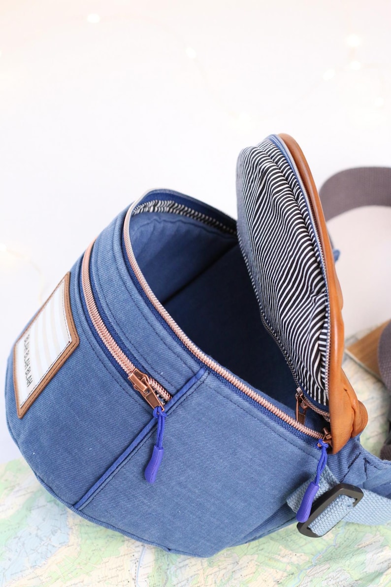 K&ouml;nnte beinhalten: Eine blaue Jeans-Bauchtasche mit einem braunen Lederriemen und einem Rei&szlig;verschluss. Das Innere der Tasche ist mit schwarz-wei&szlig; gestreiftem Stoff gef&uuml;ttert. Die Tasche hat einen kleinen Patch auf der Vorderseite mit dem Text "The Little Market" in Wei&szlig;.