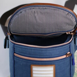K&ouml;nnte beinhalten: Eine blaue Canvas-Tasche mit einer braunen Lederklappe und einem Rei&szlig;verschluss. Die Tasche hat ein wei&szlig;-schwarz gestreiftes Futter und einen Patch mit dem Text "Blah Blah Blah" auf der Vorderseite.