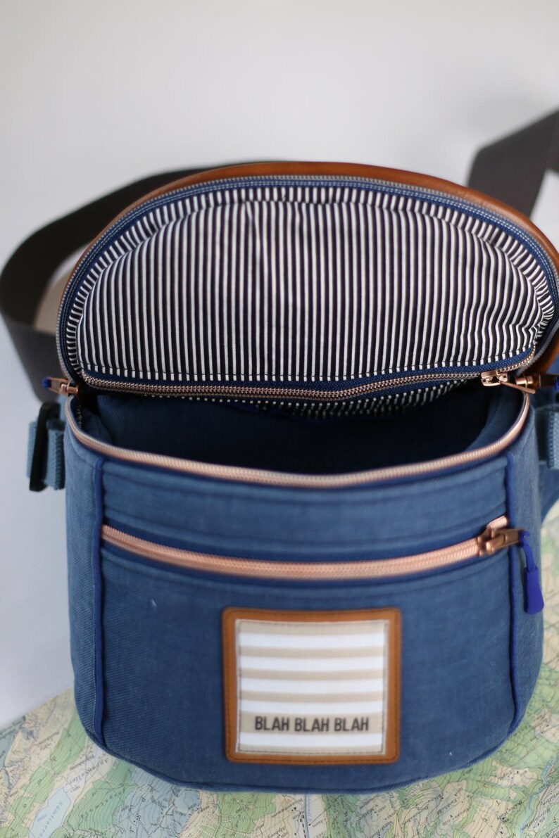 K&ouml;nnte beinhalten: Eine blaue Canvas-Tasche mit einer braunen Lederklappe und einem Rei&szlig;verschluss. Die Tasche hat ein wei&szlig;-schwarz gestreiftes Futter und einen Patch mit dem Text "Blah Blah Blah" auf der Vorderseite.
