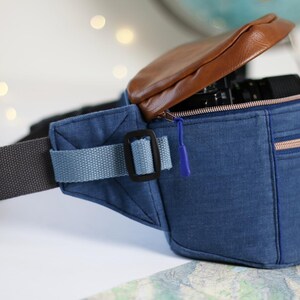 K&ouml;nnte beinhalten: Eine blaue Jeans-Bauchtasche mit einer braunen Lederklappe und einem Rei&szlig;verschluss. Die Tasche hat einen schwarzen verstellbaren Gurt und einen blauen Gurt mit einer schwarzen Schnalle. Die Tasche ist offen und eine Kamera ist im Inneren sichtbar.