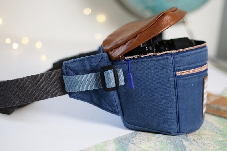 K&ouml;nnte beinhalten: Eine blaue Jeans-Bauchtasche mit einer braunen Lederklappe und einem Rei&szlig;verschluss. Die Tasche hat einen schwarzen verstellbaren Gurt und einen blauen Gurt mit einer schwarzen Schnalle. Die Tasche ist offen und eine Kamera ist im Inneren sichtbar.