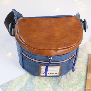 K&ouml;nnte beinhalten: Eine blaue Jeans-Umh&auml;ngetasche mit einer braunen Lederklappe und einem Rei&szlig;verschluss. Die Tasche hat ein kleines wei&szlig;es Etikett mit dem Text "Blah Blah" darauf.