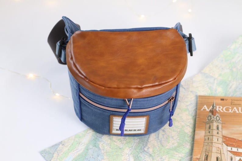 K&ouml;nnte beinhalten: Eine blaue Jeans-Umh&auml;ngetasche mit einer braunen Lederklappe und einem Rei&szlig;verschluss. Die Tasche hat ein kleines wei&szlig;es Etikett mit dem Text "Blah Blah" darauf.