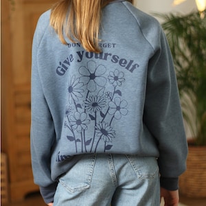 Könnte beinhalten: Ein blaues Sweatshirt mit einem Blumendesign und dem Text "Vergiss nicht, dir selbst Blumen zu schenken" auf der Rückseite.