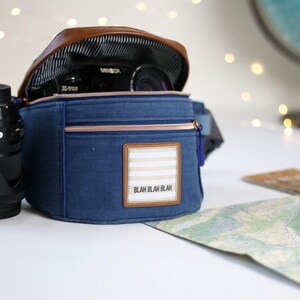 K&ouml;nnte beinhalten: Eine blaue Canvas-Kameratasche mit einer braunen Lederklappe und einem Rei&szlig;verschluss. Die Tasche hat einen wei&szlig;-braunen Streifen-Patch mit dem Text "Blah Blah Blah" darauf. Ein Kameraobjektiv ist in der Tasche sichtbar.