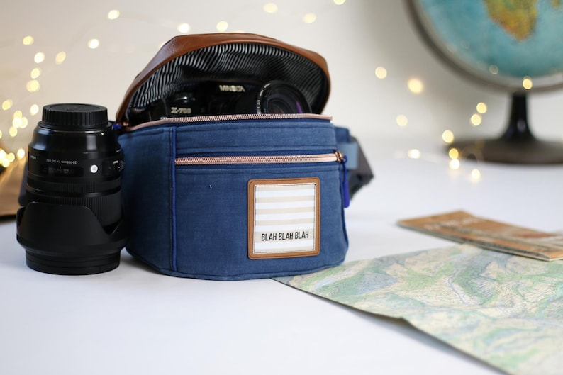 K&ouml;nnte beinhalten: Eine blaue Canvas-Kameratasche mit einer braunen Lederklappe und einem Rei&szlig;verschluss. Die Tasche hat einen wei&szlig;-braunen Streifen-Patch mit dem Text "Blah Blah Blah" darauf. Ein Kameraobjektiv ist in der Tasche sichtbar.