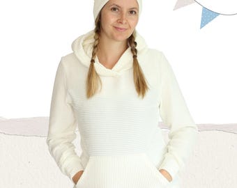 Puede incluir: Una sudadera con capucha blanca de punto de canalé con un bolsillo canguro y un cuello de pico. La sudadera la lleva puesta una persona con el pelo largo rubio y una boina blanca.