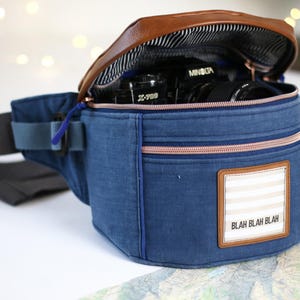 K&ouml;nnte beinhalten: Eine blaue Jeans-Bauchtasche mit einer braunen Lederklappe und einem Rei&szlig;verschluss. Die Tasche hat einen wei&szlig;-beigen Streifen-Patch mit dem Text "Blah Blah Blah" darauf. Eine Kamera ist in der Tasche sichtbar.