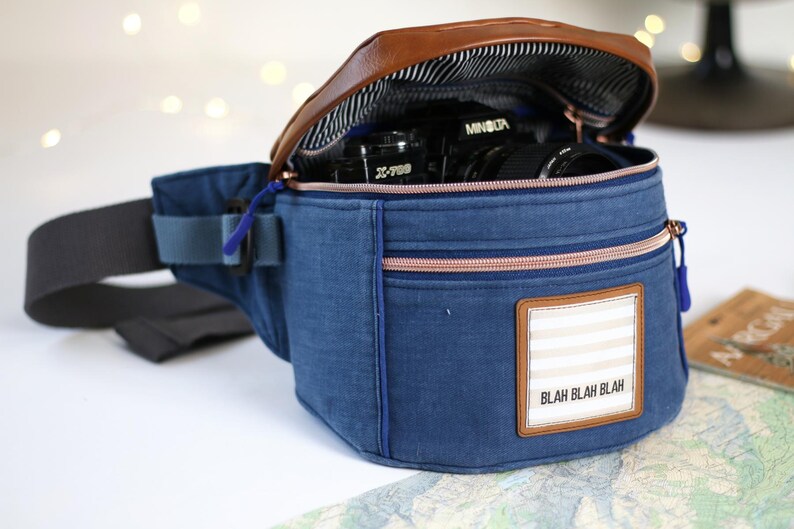 K&ouml;nnte beinhalten: Eine blaue Jeans-Bauchtasche mit einer braunen Lederklappe und einem Rei&szlig;verschluss. Die Tasche hat einen wei&szlig;-beigen Streifen-Patch mit dem Text "Blah Blah Blah" darauf. Eine Kamera ist in der Tasche sichtbar.