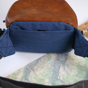 K&ouml;nnte beinhalten: Eine blaue Jeans-Kameratasche mit brauner Lederklappe und schwarzem Riemen. Die Tasche hat eine kleine Innentasche.