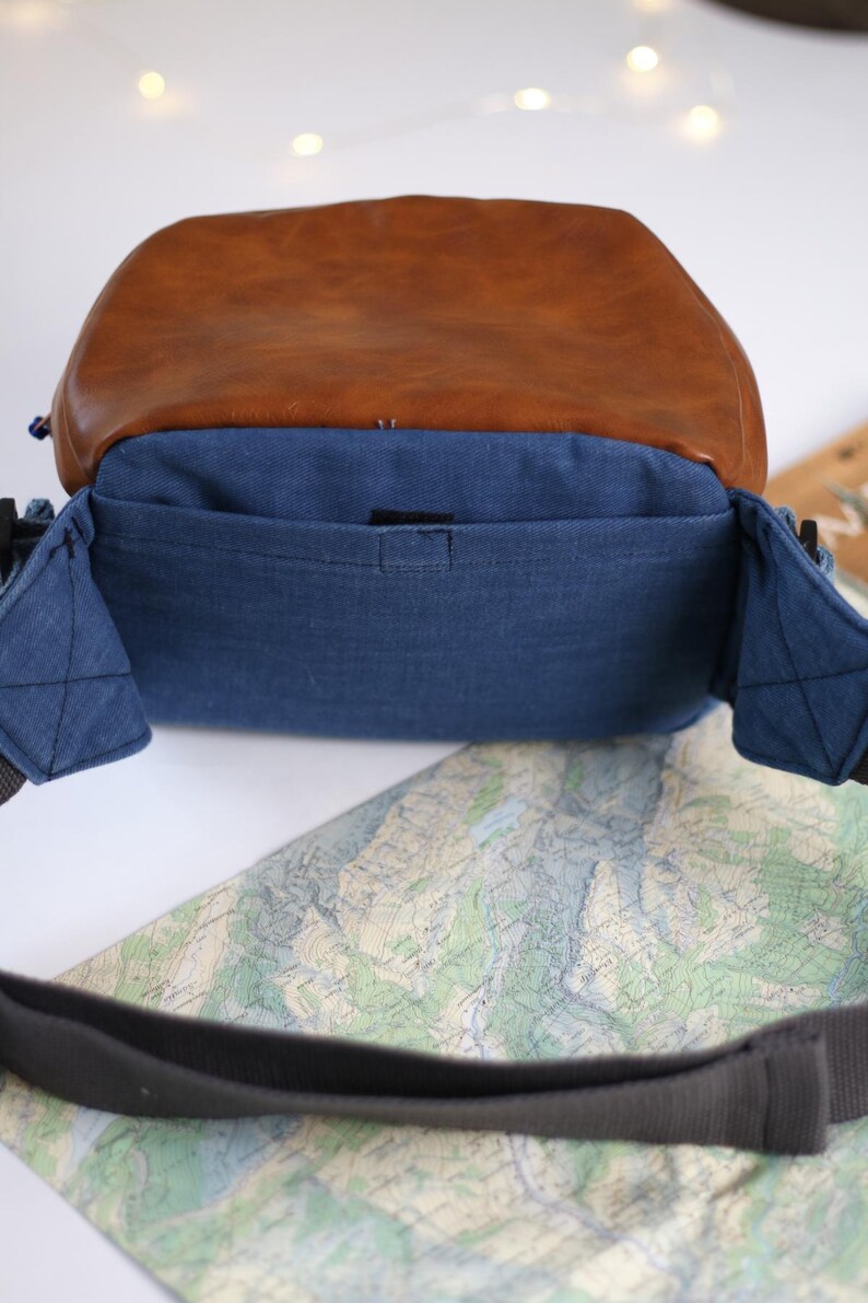 K&ouml;nnte beinhalten: Eine blaue Jeans-Kameratasche mit brauner Lederklappe und schwarzem Riemen. Die Tasche hat eine kleine Innentasche.