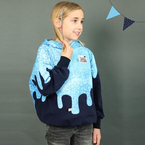 Könnte beinhalten: Ein blauer und marineblauer Hoodie mit einem tropfenden Malereimuster. Der Hoodie hat einen kleinen Patch mit einem Regenbogen und einem Herzen auf der Vorderseite.