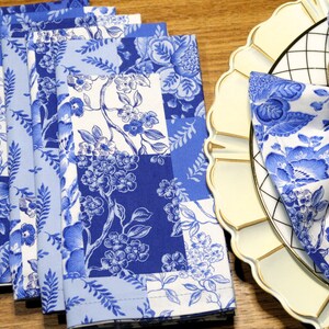 May include: A set of blue and white floral patterned cloth napkins. The napkins feature a patchwork design with varying shades of blue and white floral patterns. One napkin is folded and placed on a cream-colored plate with a gold napkin ring.