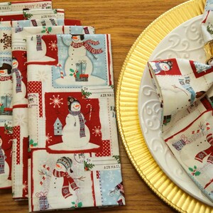 May include: A set of folded Christmas-themed cloth napkins with a snowman design. The napkins feature a red, white, and blue color scheme with snowflakes and postcard-style graphics. One napkin is folded and placed on a white plate with a gold charger.