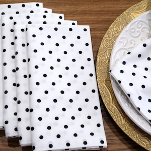 May include: White cloth napkins with black polka dots are stacked next to a gold-rimmed plate with a white napkin featuring the same polka dot pattern. A gold napkin ring holds the napkin in place.
