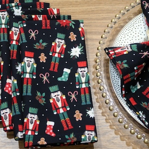 May include: A set of black Christmas-themed cloth napkins featuring a repeating pattern of nutcrackers, gingerbread men, candy canes, and stockings. One napkin is folded and placed on a clear plate with a gold beaded edge. The napkins are made of fabric.