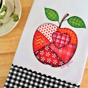 May include: A white kitchen towel with a black and white checkered border. The towel features a red apple with a heart in the center. The apple is made of patchwork fabric with different patterns, including red polka dots, red and white stripes, and red and orange zigzags. The text "ArtSandra" and "Sunny Tonini" are printed on the towel.