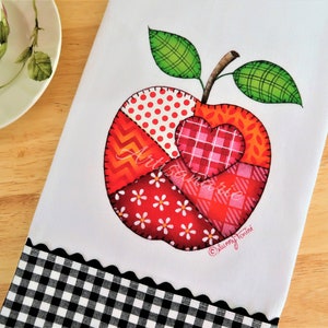 Op de afbeelding: Een witte theedoek met een rood, oranje en roze patchwork appelontwerp. De appel heeft een hart in het midden en de woorden "Art Sanatrie" staan op de appel. De theedoek heeft een zwart-wit geblokte rand.
