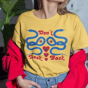May include: Mustard yellow t-shirt with a blue snake design and the text "Don't Look Back" in red. The shirt features a crew neck and short sleeves. The model is wearing a red jacket and blue and white striped jeans.
