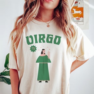 May include: Cream-colored t-shirt with the word "VIRGO" in green block letters. The shirt features a green Virgo symbol and a graphic of a person in a green dress holding a wheat stalk. The shirt is a casual style.