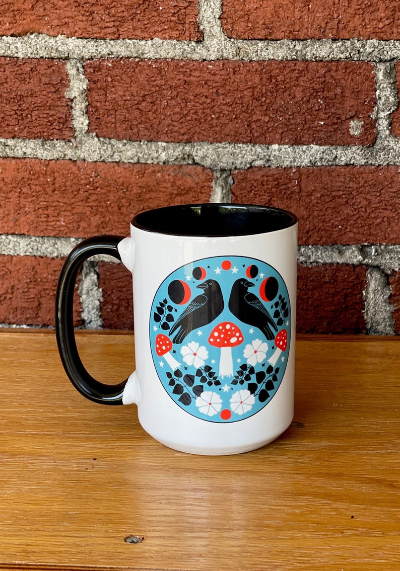 Puede incluir: Taza de cer&aacute;mica blanca con asa e interior negros. La taza presenta un dise&ntilde;o circular azul con dos cuervos negros, setas rojas, flores blancas y follaje negro. El fondo es una pared de ladrillo rojo.