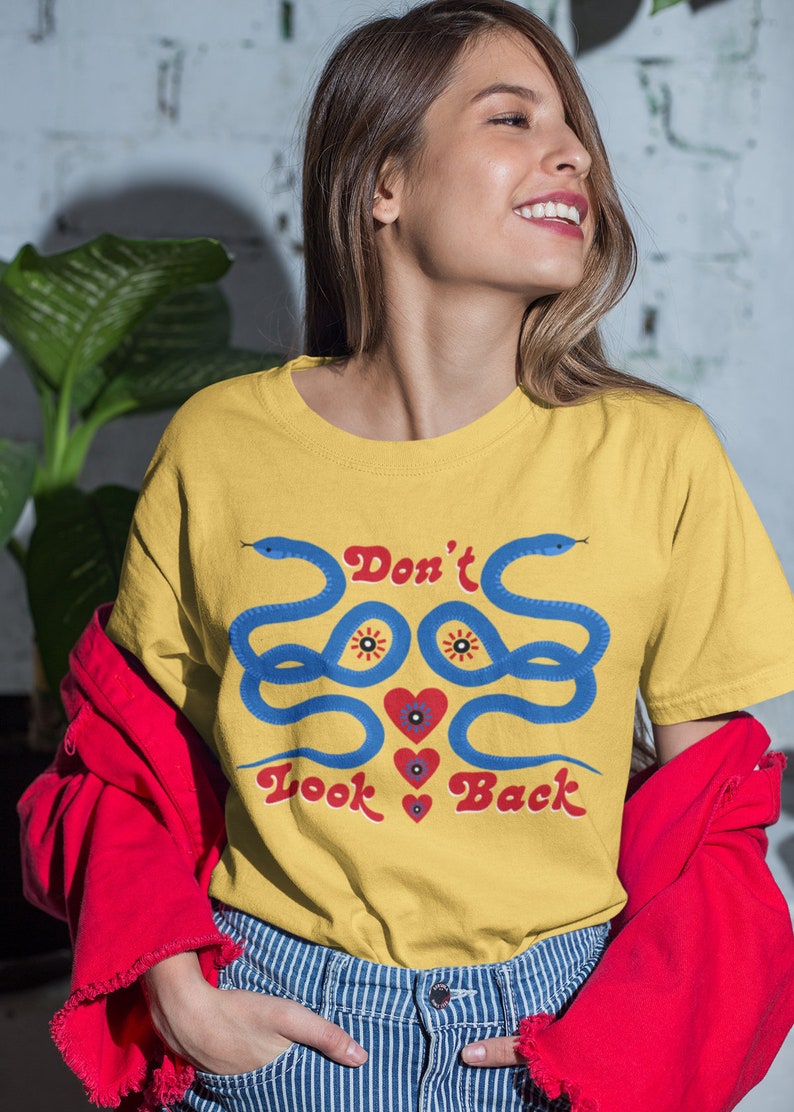 May include: Mustard yellow t-shirt with a blue snake design and the text "Don't Look Back" in red. The shirt features a crew neck and short sleeves. The model is wearing a red jacket and blue and white striped jeans.