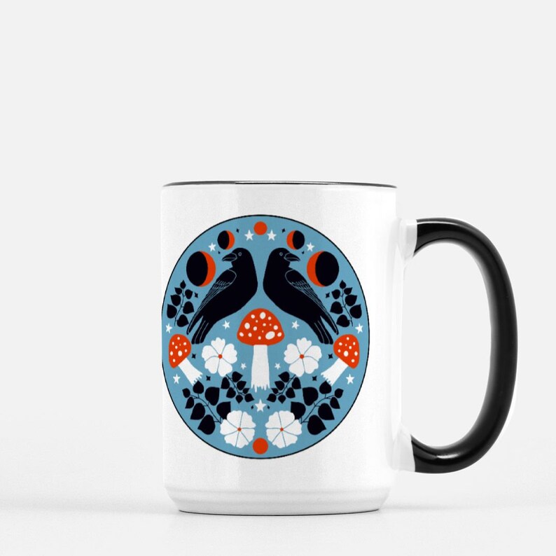 Puede incluir: Taza de cer&aacute;mica blanca con asa y borde negros. La taza presenta un dise&ntilde;o circular con dos cuervos negros, setas rojas y blancas, flores blancas y lunas crecientes sobre un fondo azul.
