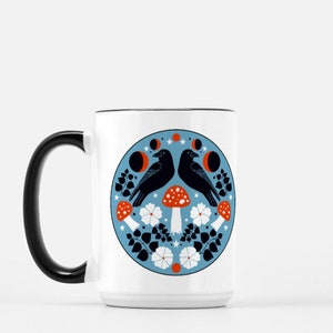 Puede incluir: Taza de cer&aacute;mica blanca con asa y borde negros. La taza presenta un dise&ntilde;o circular con dos cuervos negros, setas rojas y blancas, flores blancas y lunas crecientes sobre un fondo azul claro. El dise&ntilde;o est&aacute; centrado en la taza.