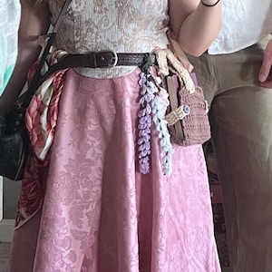 May include: A pink skirt with a floral pattern, a brown leather belt, and a crocheted flower garland. The skirt is worn with a white top and a brown leather bag.