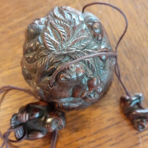 May include: A dark brown, intricately carved wooden bead with a frog motif. The bead is round with detailed carvings of frogs and foliage. Two smaller frog-shaped beads are attached with brown cords.