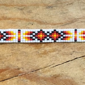 May include: White beaded bracelet with a geometric pattern in red, orange, and black. The bracelet has silver clasps.