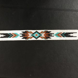 K&ouml;nnte beinhalten: Ein wei&szlig;es Perlenarmband mit einem geometrischen Muster in Schwarz, Braun und T&uuml;rkis.