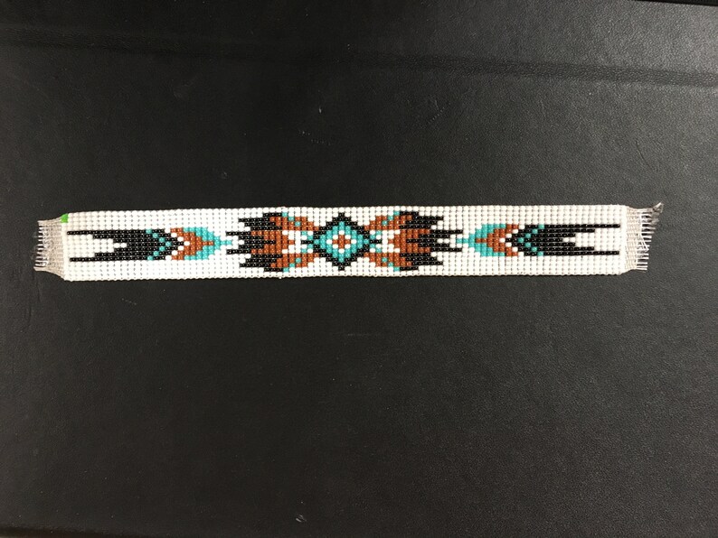 K&ouml;nnte beinhalten: Ein wei&szlig;es Perlenarmband mit einem geometrischen Muster in Schwarz, Braun und T&uuml;rkis.