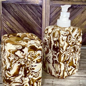 Peut inclure: Un ensemble de salle de bain assorti comprenant un distributeur de savon avec une pompe blanche et un gobelet. Les deux articles présentent un motif marbré dans les tons marron et crème. L'ensemble est présenté sur une surface en bois.