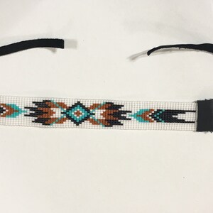 K&ouml;nnte beinhalten: Ein wei&szlig;es Perlenhaarband mit einem geometrischen Muster in Schwarz, Braun und T&uuml;rkis. Das Haarband hat an jedem Ende schwarze Lederb&auml;nder.
