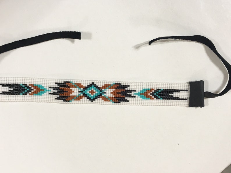 K&ouml;nnte beinhalten: Ein wei&szlig;es Perlenhaarband mit einem geometrischen Muster in Schwarz, Braun und T&uuml;rkis. Das Haarband hat an jedem Ende schwarze Lederb&auml;nder.
