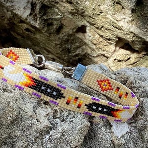 May include: A beaded bracelet with a geometric pattern in shades of beige, black, white, orange, and purple. The bracelet has a silver clasp.