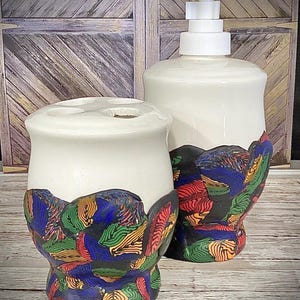Peut inclure: Ensemble de distributeur de savon et de porte-brosse à dents en céramique blanche. La base de chaque pièce présente un motif abstrait coloré avec des touches de rouge, vert, bleu et jaune. Le distributeur a une pompe blanche.