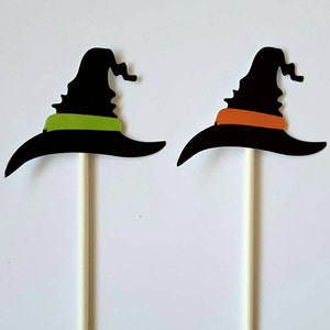 May include: Two black witch hat cake toppers with green and orange trim. The hats are attached to white sticks.