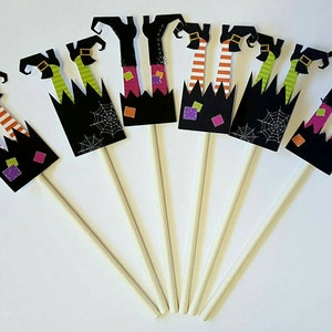 May include: Set of eight witch leg cupcake toppers. Each topper features black witch legs with striped stockings, a black witch hat, and a black and green witch skirt. The toppers are attached to wooden sticks.