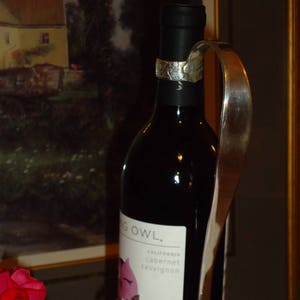 May include: A silver bottle stopper with a curved handle, holding a bottle of red wine. The wine bottle label reads "Flying Owl. California Cabernet Sauvignon".