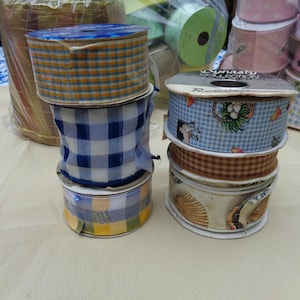 May include: Two stacks of assorted craft ribbon spools featuring various patterns including blue and yellow plaid, navy and white gingham, a light blue print with small illustrations, a brown checkered pattern, and a seashell print design.