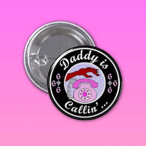 May include: A black and white button with a pink rotary phone and a red hand reaching for it. The text "Daddy is Callin' ..." is written around the phone and hand. The background is a glittery silver.