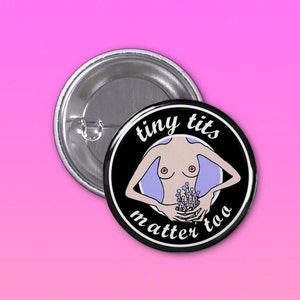Może przedstawiać: Czarny, okrągły guzik z białym napisem "tiny tits matter too". Guzik przedstawia ilustrację torsu z ramionami i małymi piersiami, trzymającego kwiaty. Guzik leży obok srebrnej agrafki.