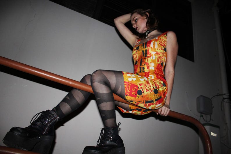 May include: A sleeveless, bodycon dress with a vibrant orange and yellow background, featuring a collage of text and graphics. The outfit is completed with black platform boots and striped tights. The model is sitting on a brown metal bar.