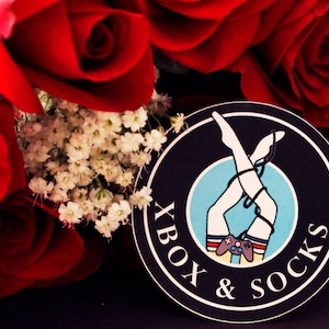 Op de afbeelding: Een zwart-witte sticker met de tekst "Xbox & Socks" en een afbeelding van een vrouw met witte sokken en rood-wit-blauwe strepen om haar dijen. De vrouw houdt een videogamecontroller vast. De sticker is op een achtergrond van rode rozen.